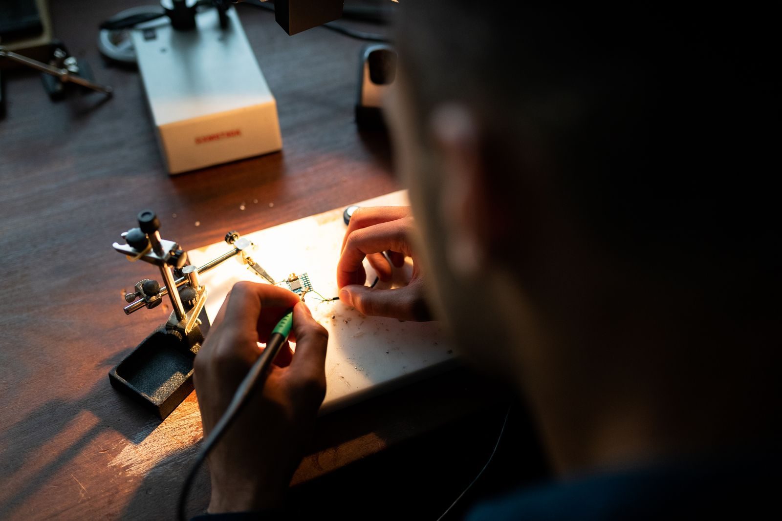 A mechanical engineer soldering a prototype circuit board at a workbench under warm lamplight.